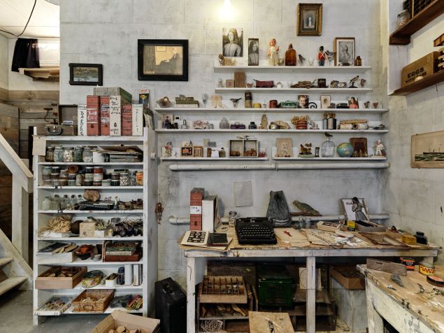 A cluttered workspace with a table and many shelves crammed with objects of all kinds