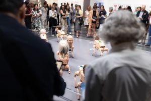 A crowd of visitors gathers around an enclosure where several small four-legged robot creatures with realistic human heads move across the floor while people photograph and watch them.