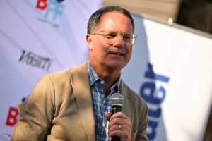Man in beige blazer stands outside holding microphone