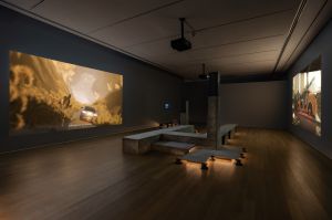 A gallery with multiple large-scale projections showing animated scenes from Lawrence Lek’s NOX series. In the center sits a modular concrete-like pavilion of tiled benches and vertical supports, lit from below with warm LEDs that accent the structure.