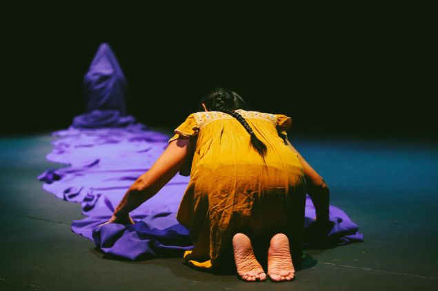 A barefoot woman in a yellow tunic dress sits on her knees before the end of massive train of blue cloth. The cloth is worn by a figure who sits several meters away from her in the background. The woman in yellow bends over the blue cloth as though folding it.