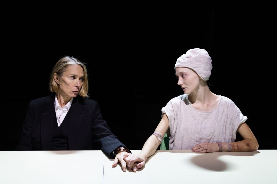 Elizabeth Marvel and another actor sit across a table holding hands, with Marvel looking gravely toward her scene partner, conveying a moment of fragile connection within the play’s otherwise cold system.