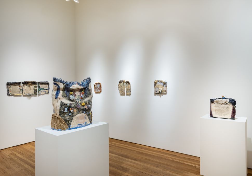 A gallery installation view shows multiple ceramic works arranged on the walls and two large ceramic sculptures on white pedestals, all illuminated by overhead spotlights on wooden floors.If you'd prefer these written in your Observer alt-text style (more concise, more atmosphe