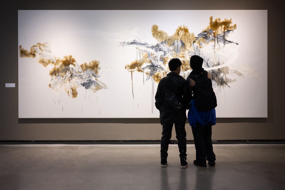 A gallery installation view showing two visitors standing in front of a large white canvas with gold and gray brushwork, emphasizing the scale and texture of Shen Wei’s recent paintings.