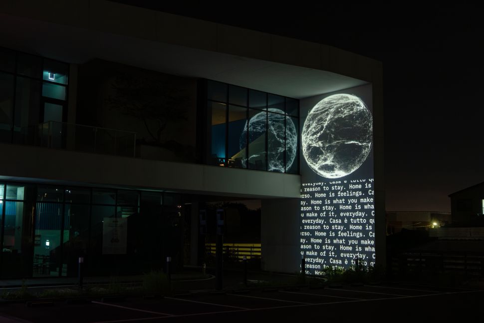 A nighttime projection artwork illuminates the corner of a modern building, showing two glowing, white, spherical forms that resemble abstract planets beside scrolling text about “home.” The projection reflects in the building’s glass windows, casting soft light into the dark, empty parking lot below.